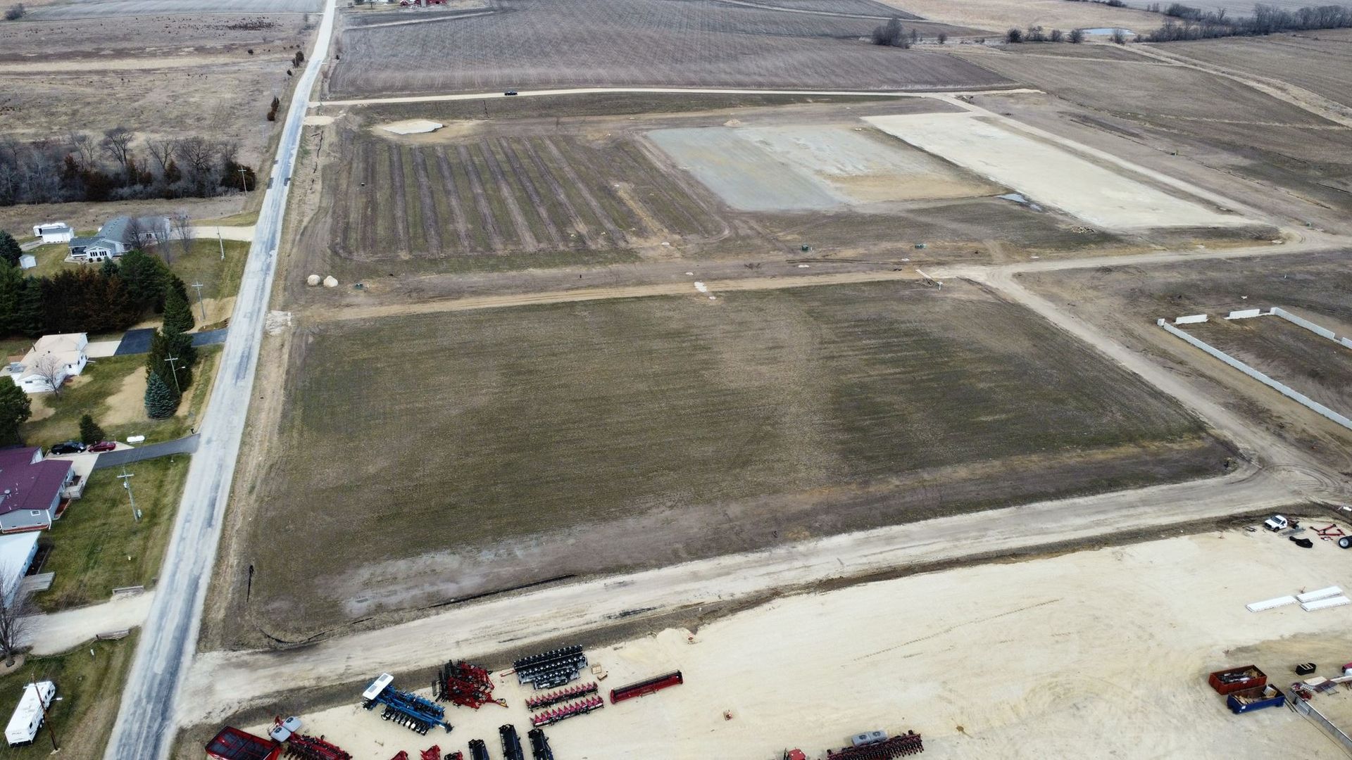 Aerial view of large plowed farmland with roads, scattered buildings, and construction equipment near the bottom edge