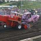 Two battered race cars, red and purple, collide on a dirt track as spectators watch nearby.