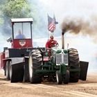 Green tractor pulling a heavy sled, kicking up dust on a dirt track with an American flag on top