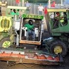 Front view of a green tractor with a snowplow blade, operator in cab, at an outdoor event