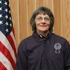 Woman standing indoors before an American flag, wearing a dark jacket with a logo.