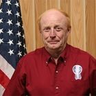 Man in a red shirt standing before an American flag and wood-paneled wall