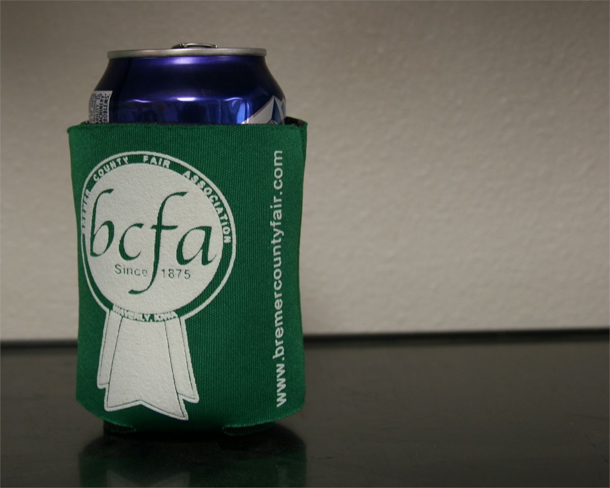 Green koozie holding a blue can with white “bcfa” logo on a dark tabletop