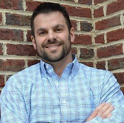 READ BIO Man in blue plaid shirt smiles, arms crossed, in front of a brick wall.