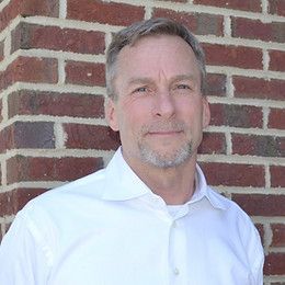 Man in white shirt against a brick wall, smiling slightly.