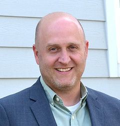 Bald man in a blue blazer and green shirt smiles broadly in front of a light blue building.