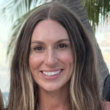 Woman with long, wavy brown hair smiles at the camera, blurred tropical background.