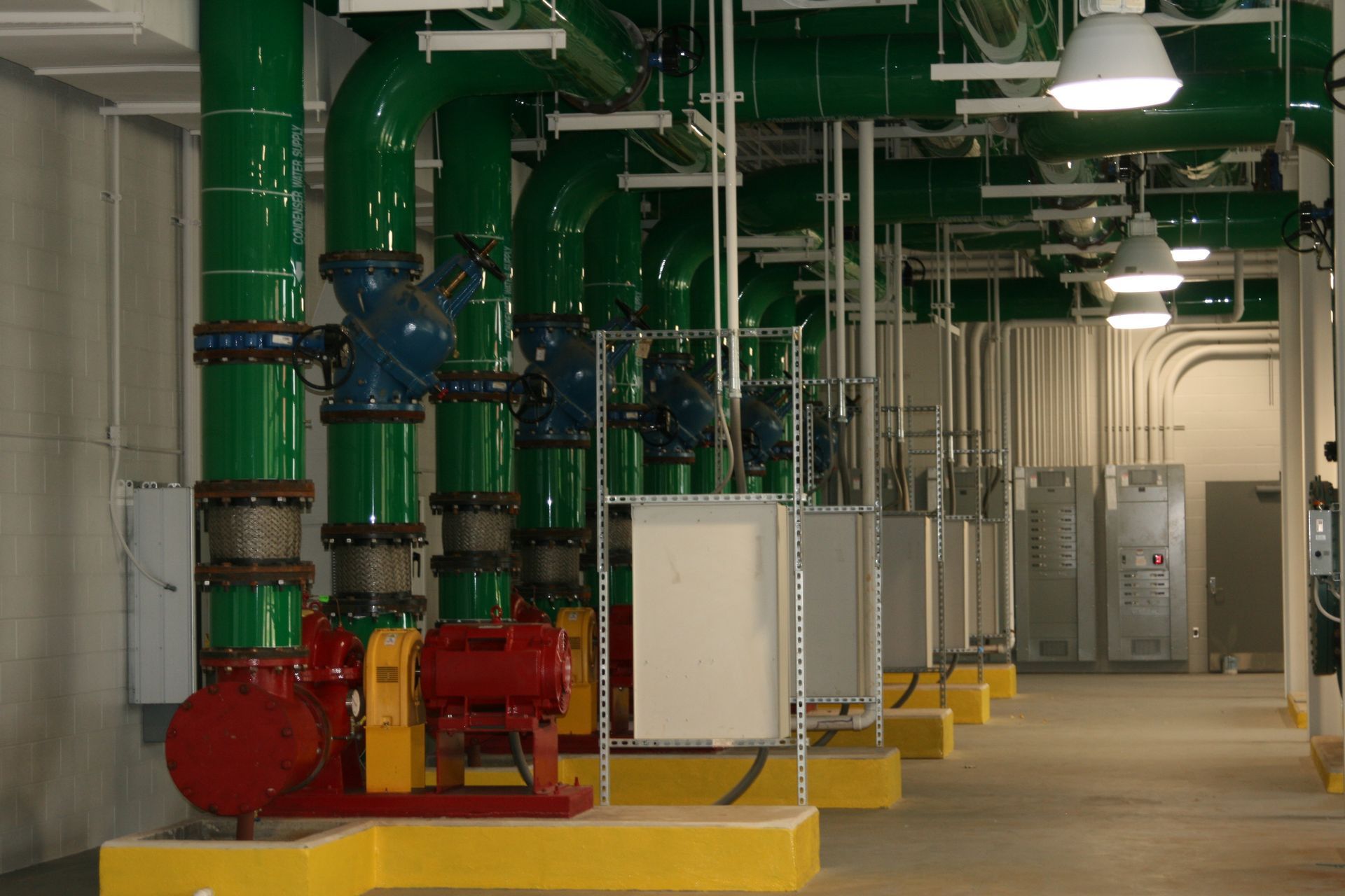 Industrial mechanical room with green pipes, valves, and electrical equipment on a concrete floor.