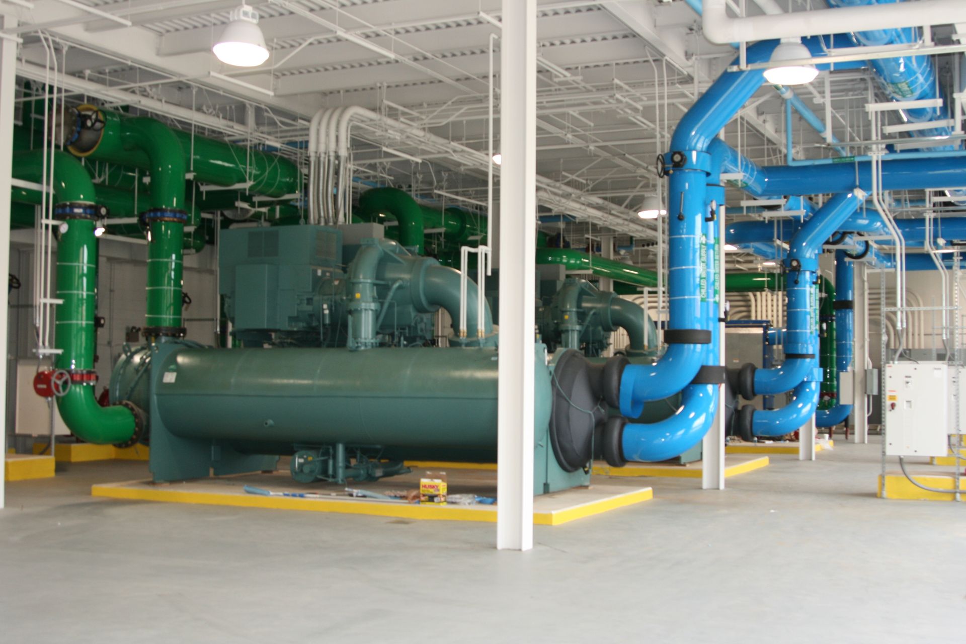 Industrial plant room with large green and blue pipes and machinery on a concrete floor