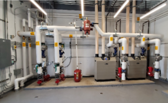 Industrial mechanical room with large white pipes, valves, and red pumps along a concrete floor.