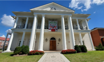 White-columned building with a central banner, red flowers, and a walkway under a blue sky