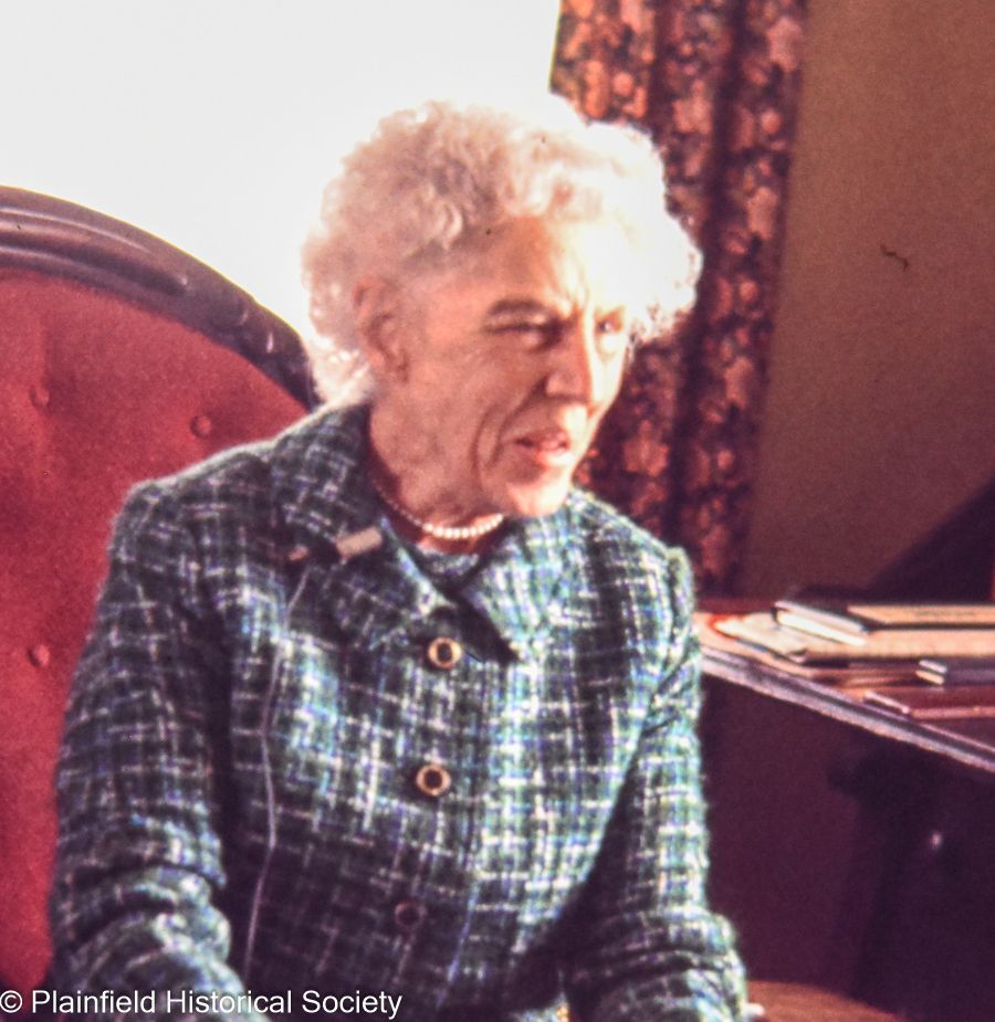 An elderly woman in a plaid jacket sits in a chair