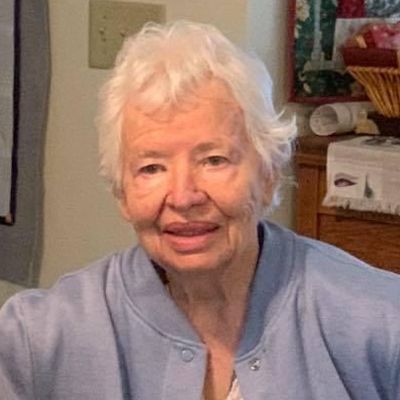 An elderly woman with white hair is wearing a blue sweater and smiling for the camera.