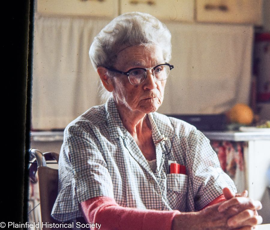 An elderly woman wearing glasses sits at a table with her hands folded