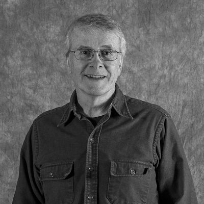 A man wearing glasses and a black shirt is smiling in a black and white photo.
