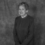 An elderly woman is standing in front of a black background in a black and white photo.