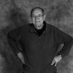 A black and white photo of an older man standing with his hands on his hips.