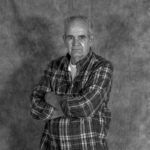 A man in a plaid shirt is standing with his arms crossed in a black and white photo.