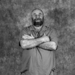 A man with a beard is standing with his arms crossed in a black and white photo.
