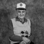 An elderly man wearing a hat and vest is sitting in a chair.