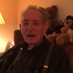 An elderly man wearing suspenders is sitting in a chair