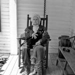 An elderly woman is sitting in a rocking chair on a porch holding a cat.