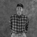 A man in a plaid shirt and hat is smiling in a black and white photo.