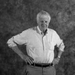A black and white photo of an older man standing with his hands on his hips.