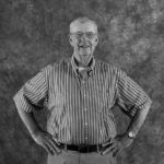 A man in a striped shirt is standing with his hands on his hips in a black and white photo.