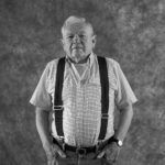A black and white photo of an older man wearing suspenders and a plaid shirt.