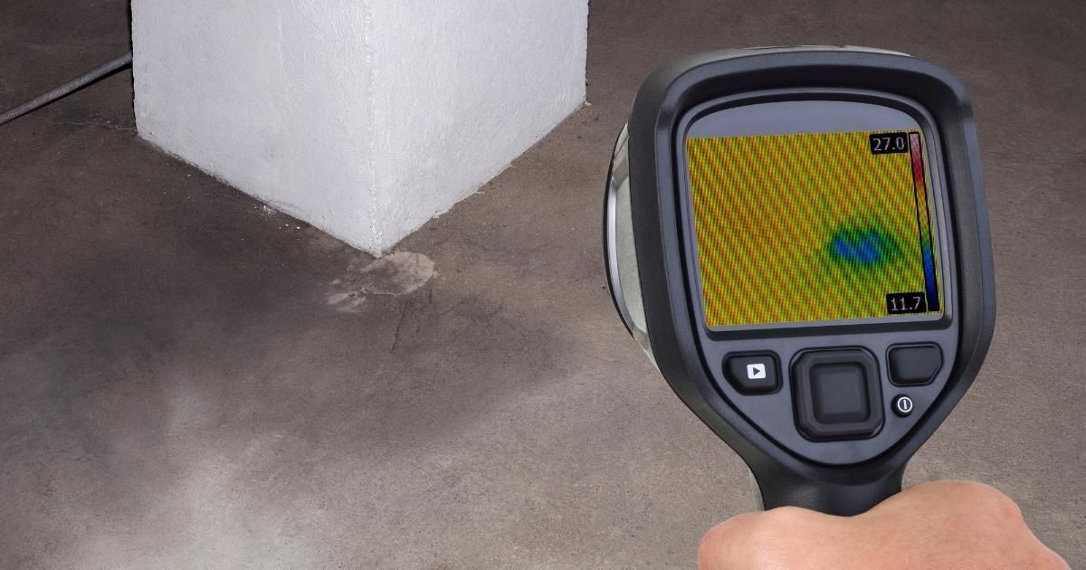 Handheld thermal camera aimed at a wall corner with a visible heat pattern on the screen