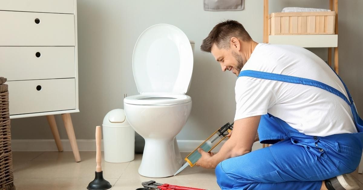 Plumber applying caulk to a toilet in a bathroom, wearing blue overalls.