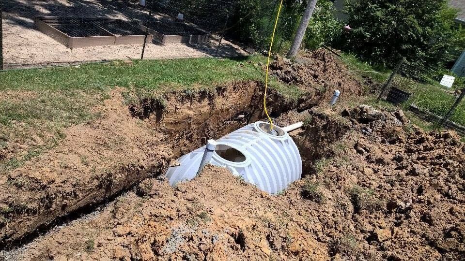 Collapsed white culvert pipe exposed in a muddy roadside trench beside grass and trees