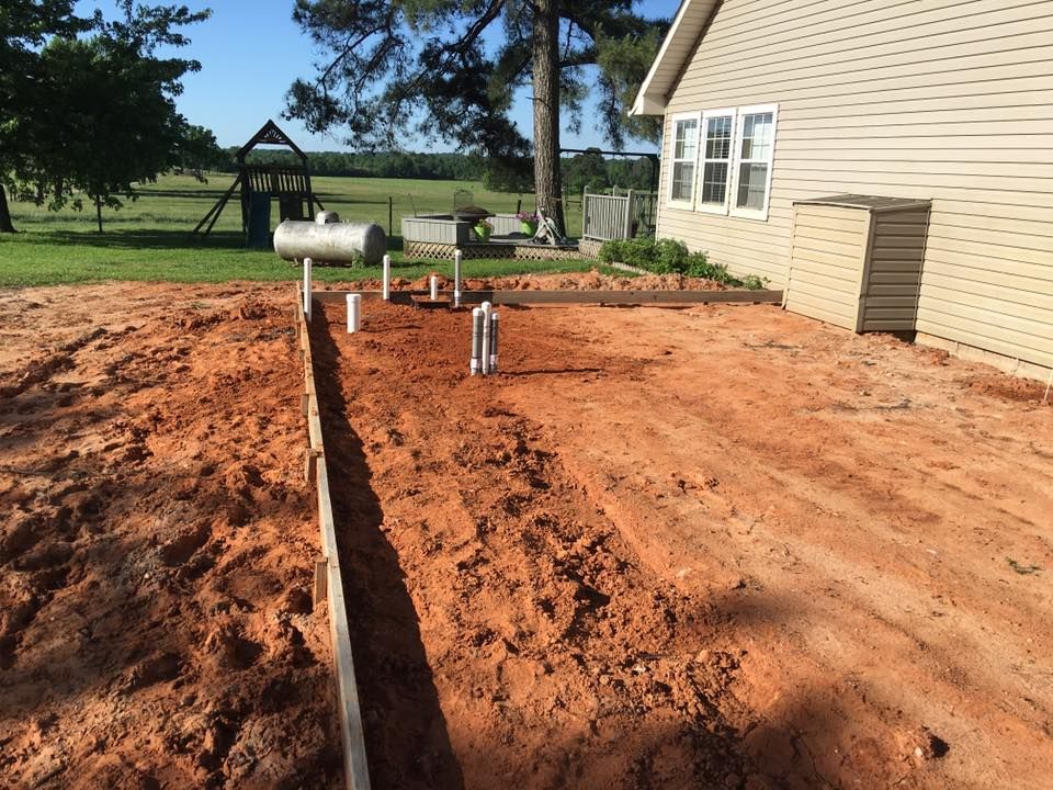 Red clay trench and utility pipes beside a beige house, with a metal tank in a yard.