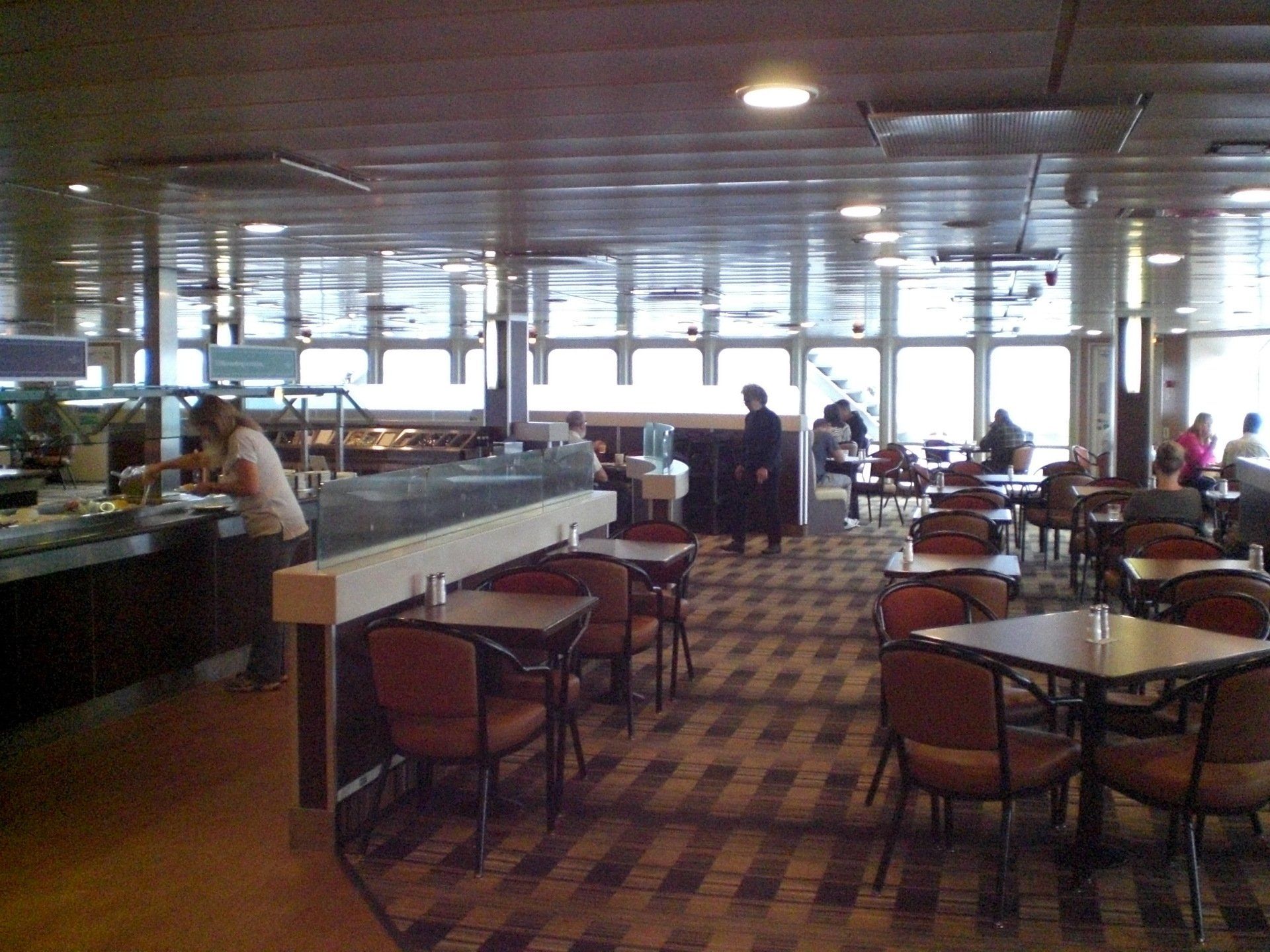 Cafeteria interior with people, tables, chairs, and a food serving area, windows with sunlight and a patterned carpet.