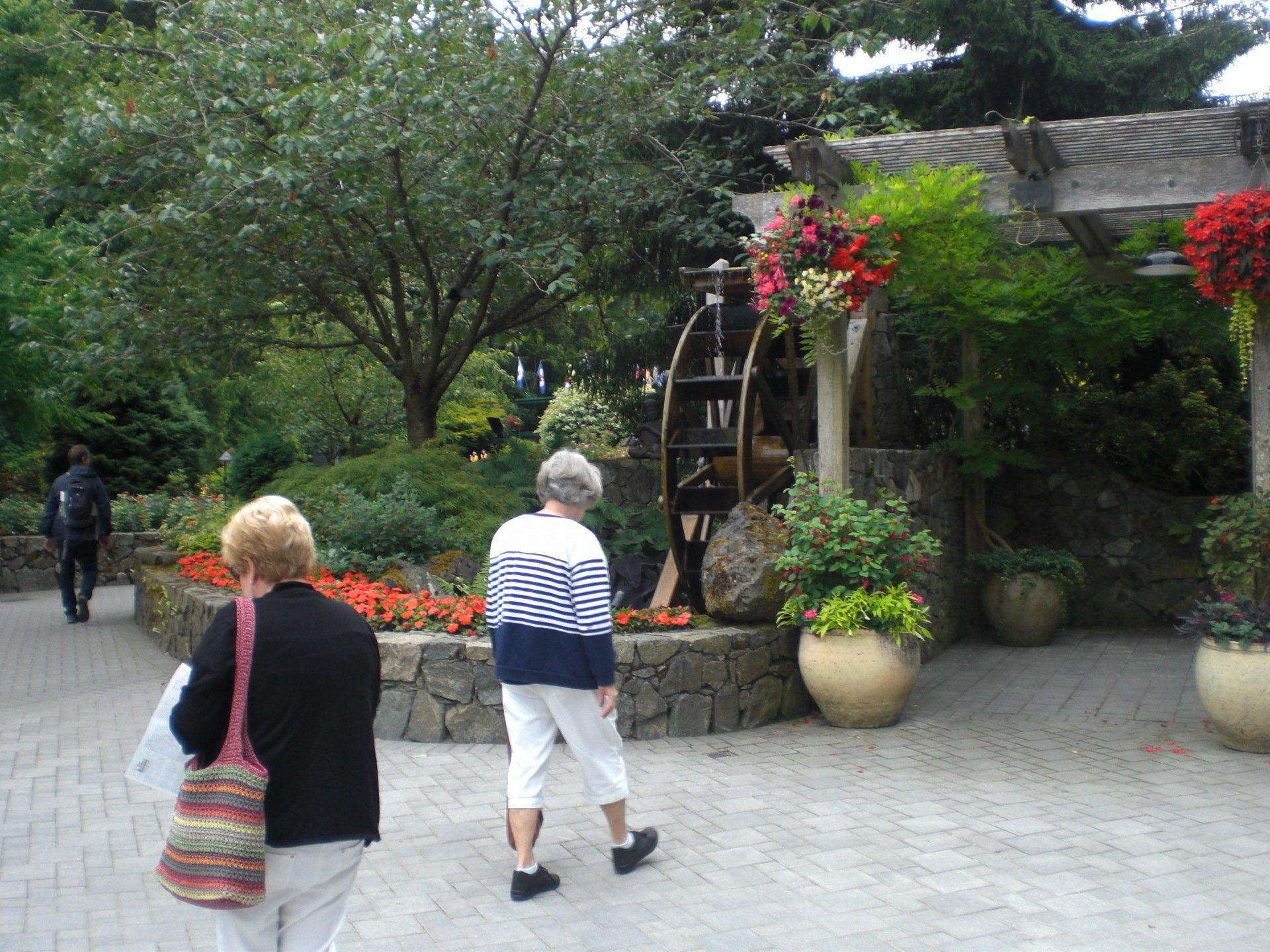 Two women walk past a water wheel and flower display in a garden.