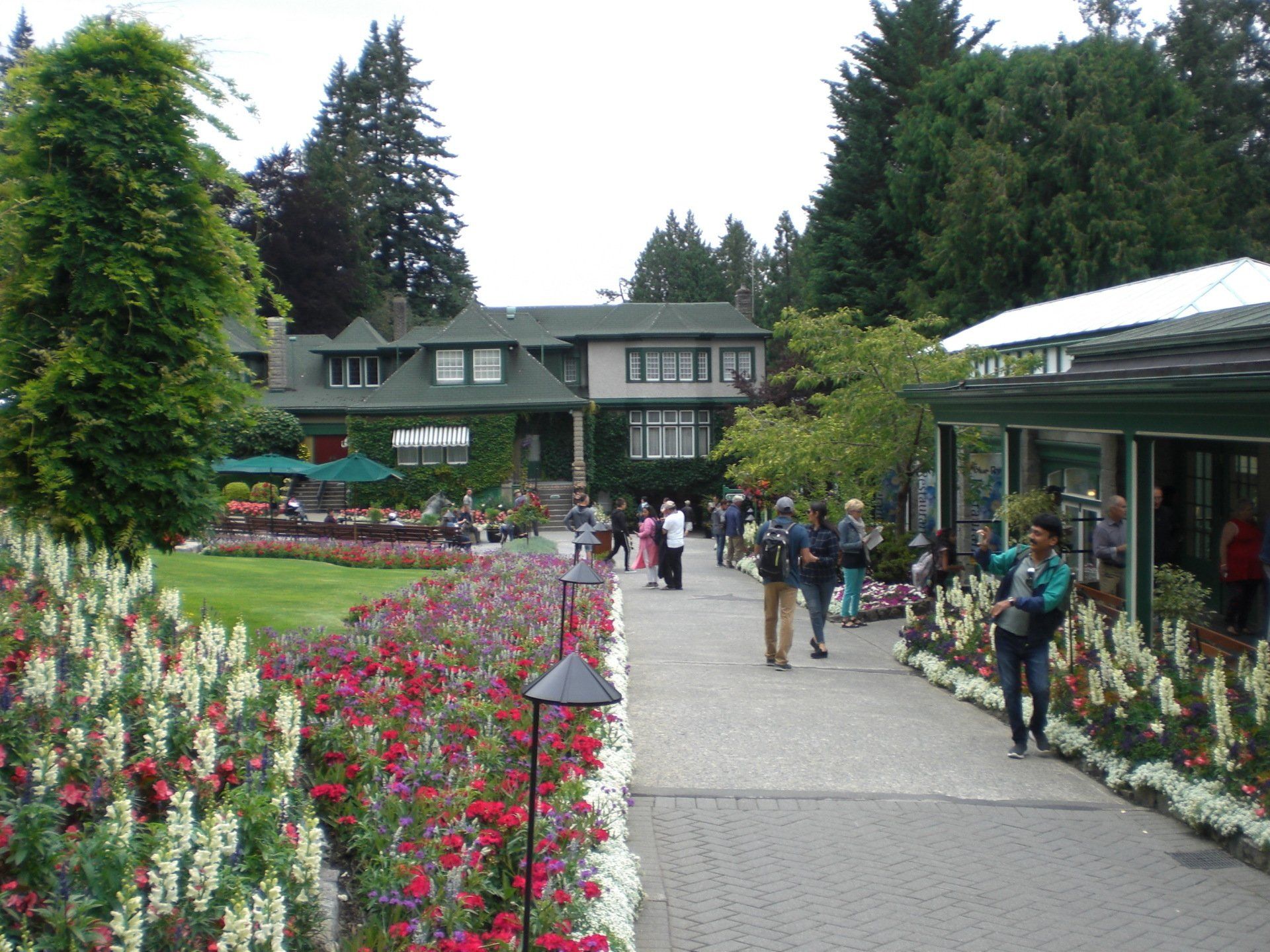 Stone walkway lined with colorful flowers and people, leading toward a large building with ivy and green roof.