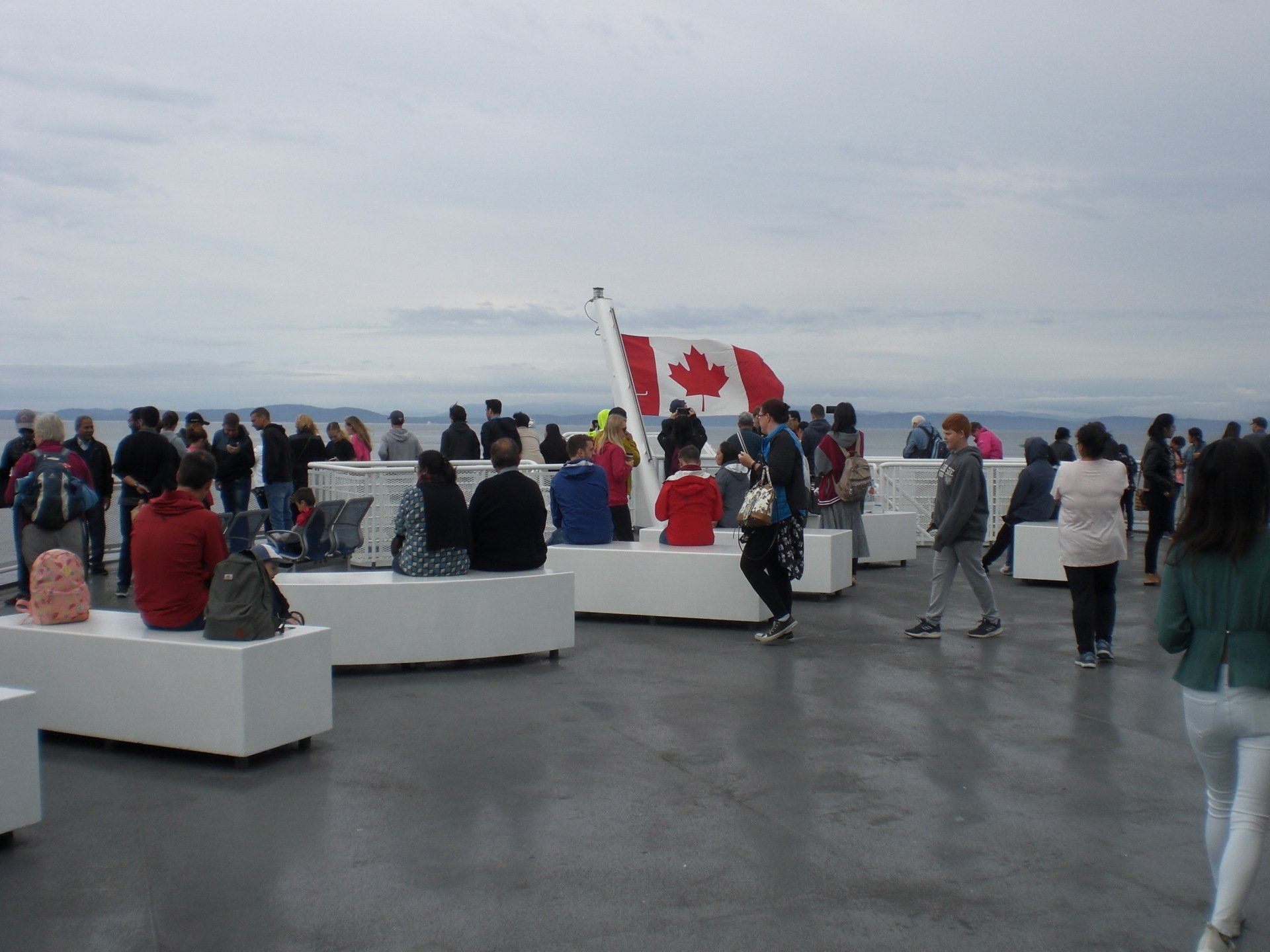 People on a ferry deck with Canadian flag. Cloudy sky, white benches, and passengers of various ages.