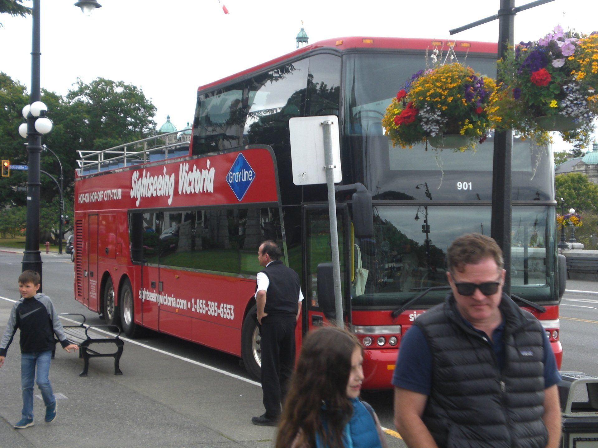 Red double-decker Sightseeing Victoria bus parked on a city street. People wait nearby, flowers hang above.