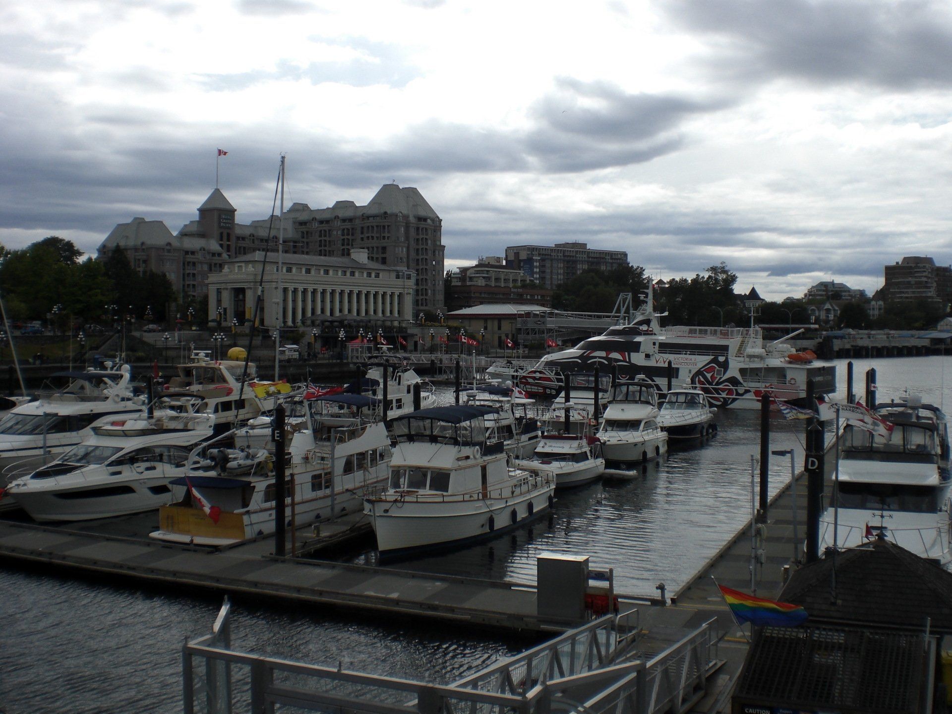 Boats docked in a harbor, with a large building in the background under an overcast sky.
