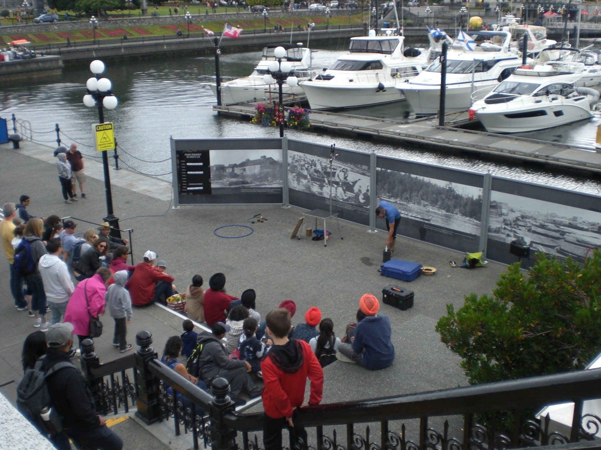 A street performer entertains a crowd near a marina, with boats docked in the background.