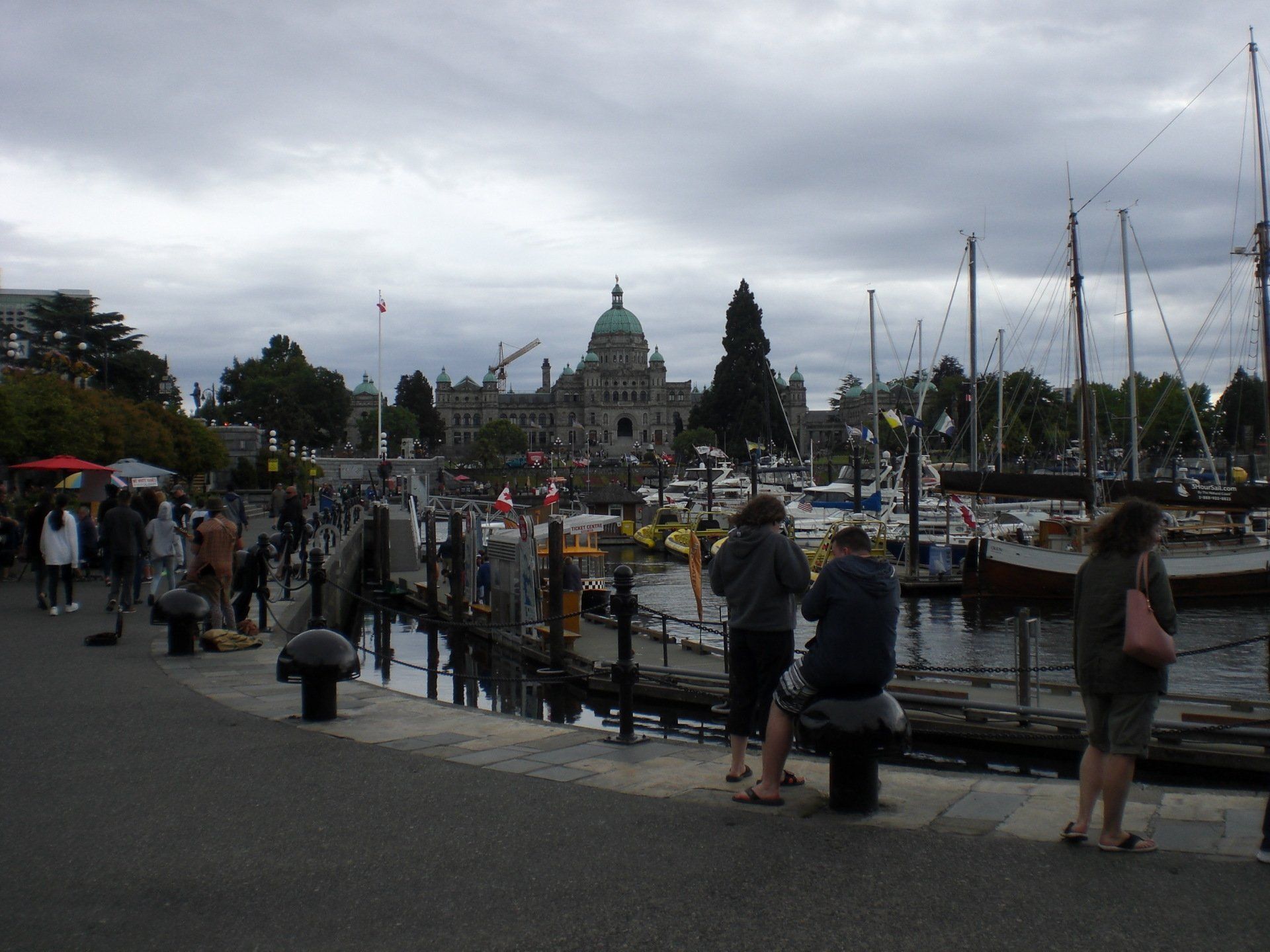 People near a harbor, the Parliament Buildings in the distance. Cloudy sky, boats, and a boardwalk are visible.