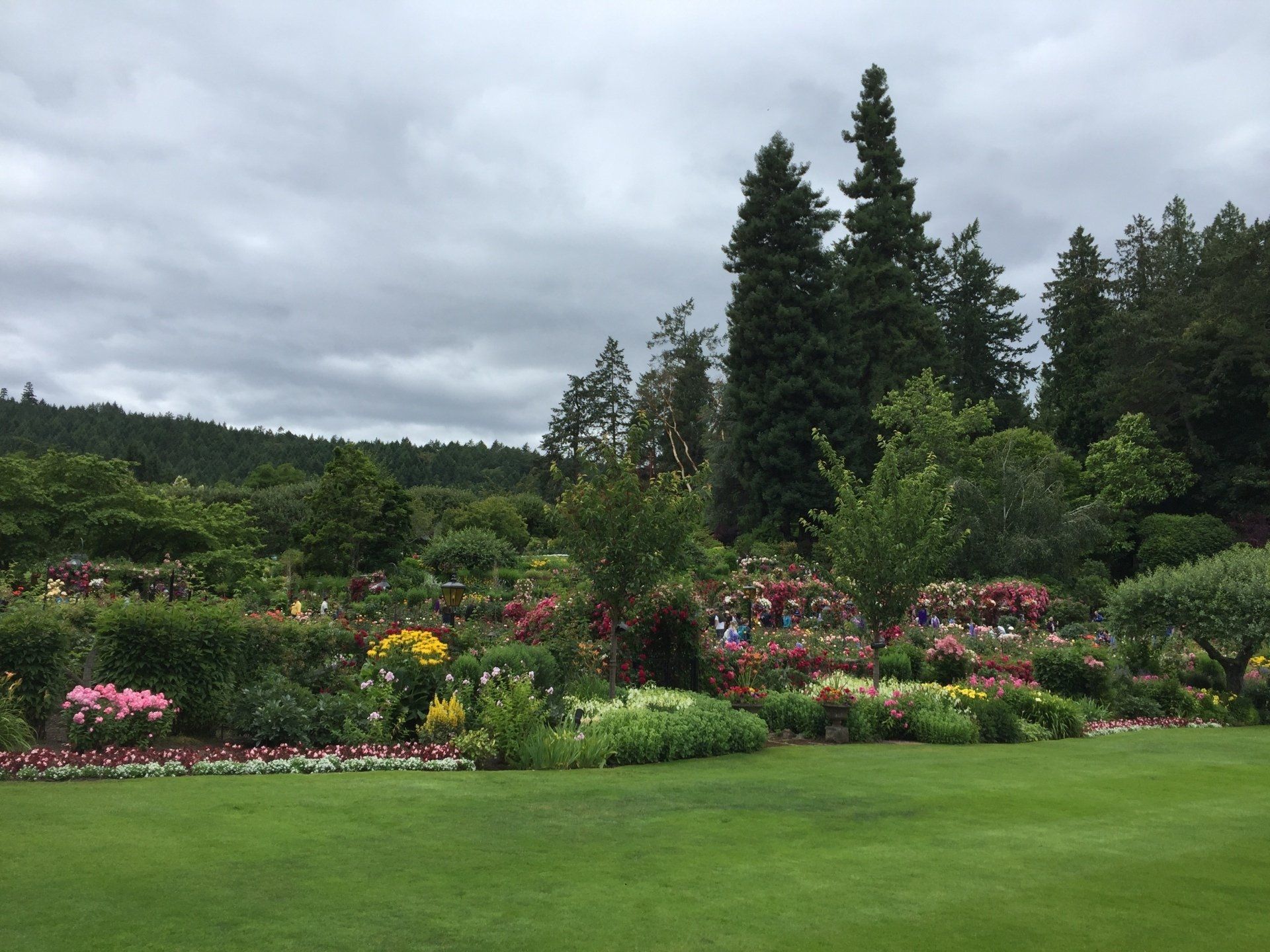 Lush garden with colorful flowers, green lawn, and tall trees under a cloudy sky.