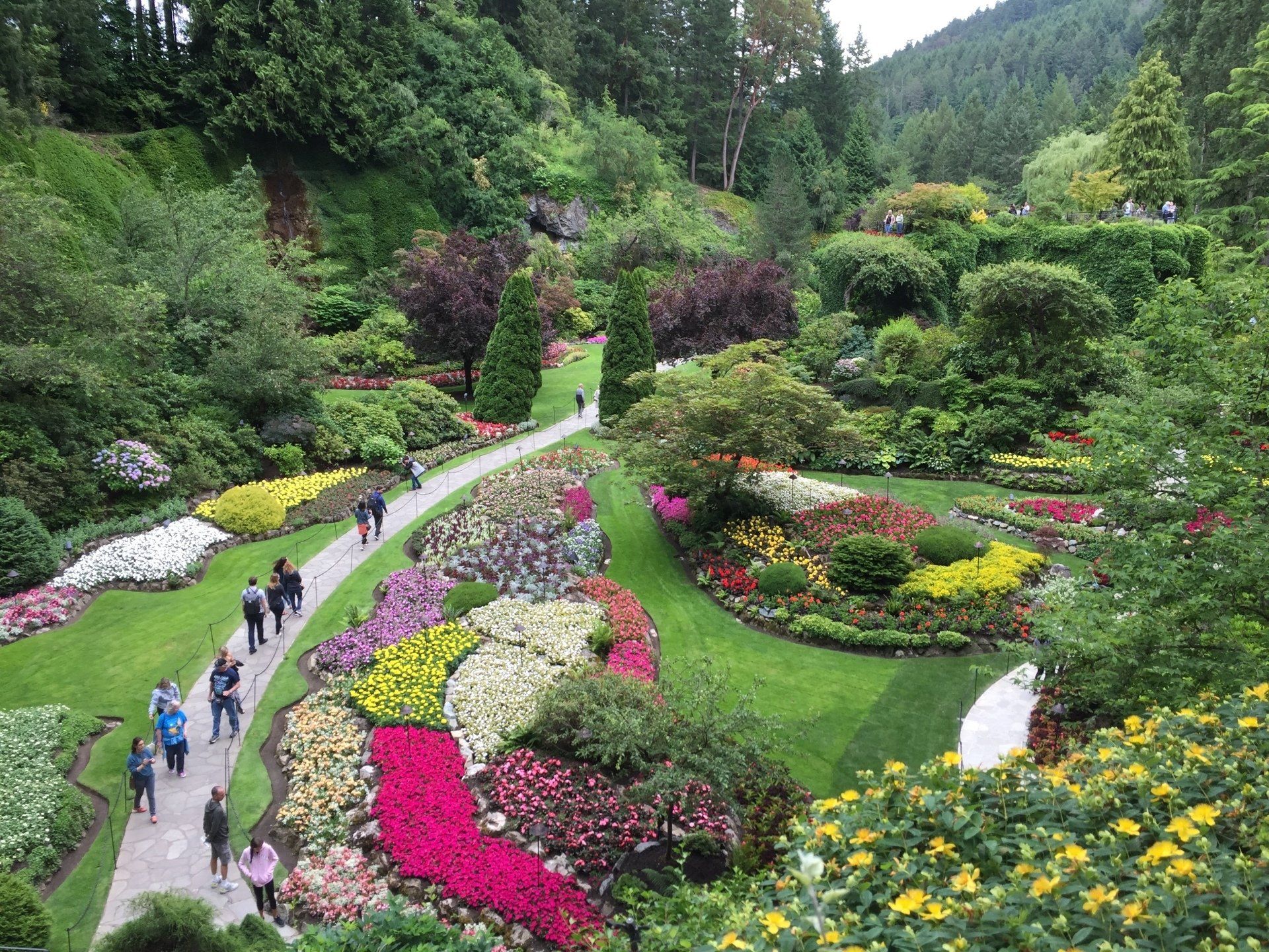 Lush garden with vibrant flowerbeds and walking paths. People stroll through the scenic landscape.