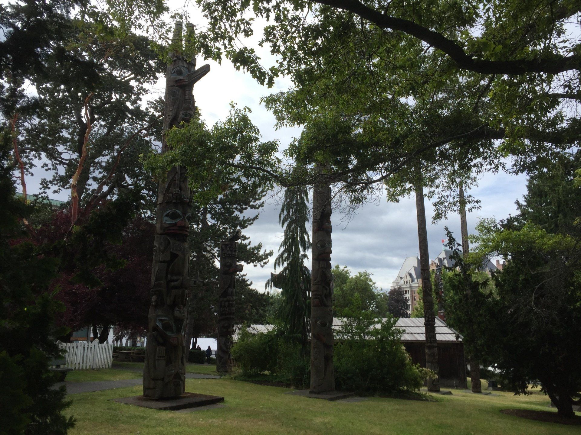 Totem poles stand in a grassy park with trees, overcast sky, and glimpse of water.