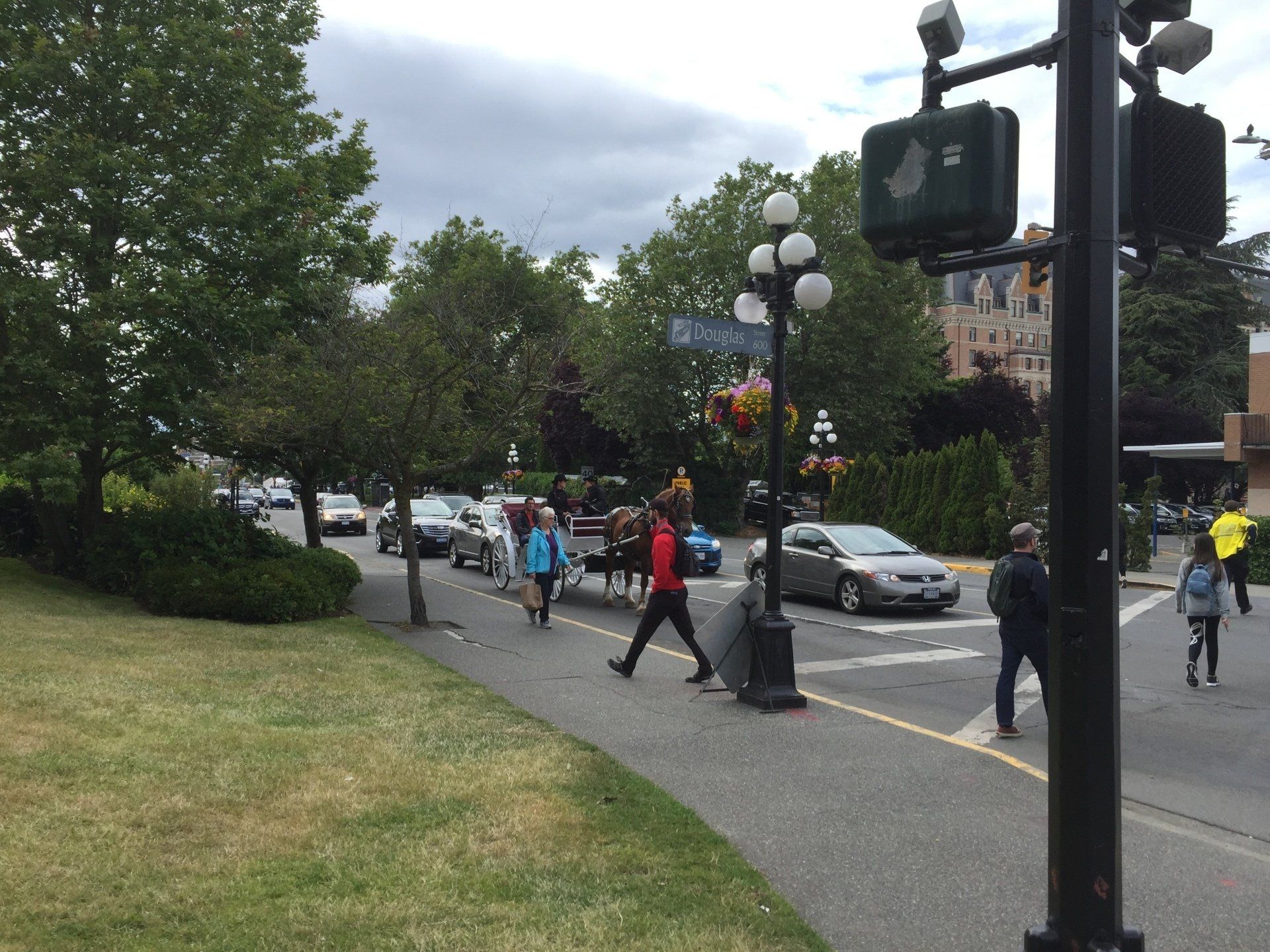 People crossing a street near a park with cars, trees, and a horse-drawn carriage. Cloudy day.