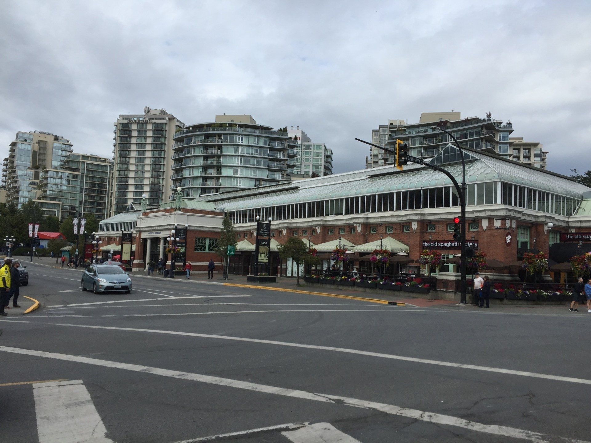 Street scene with a building, shops, cars, pedestrians, and tall apartments under cloudy skies.