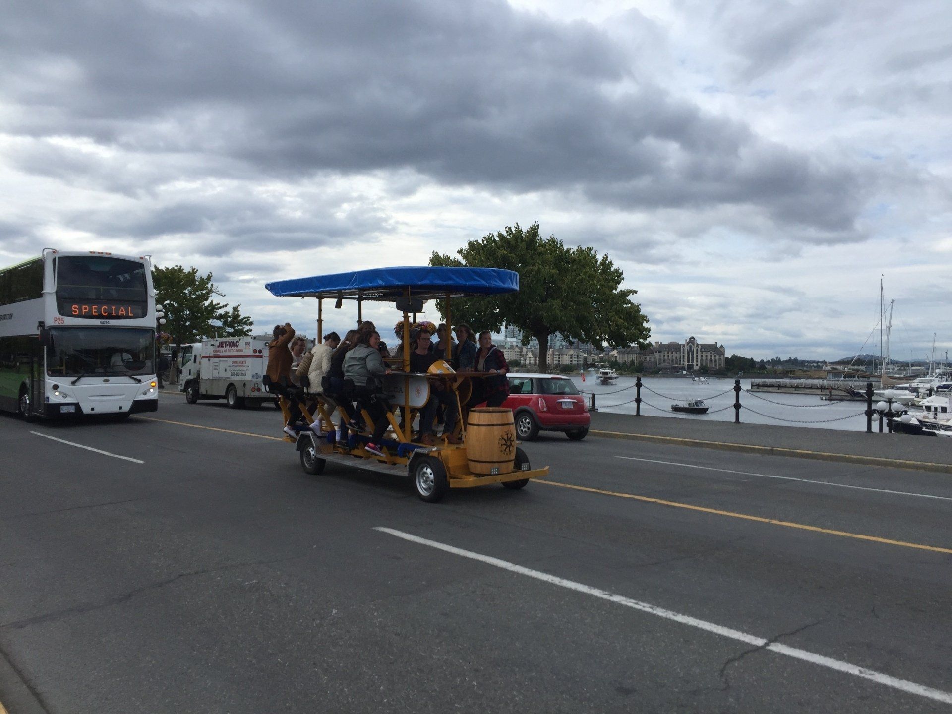 A group of people on a pedal-powered party bike traveling on a road next to water, with a double-decker bus nearby.