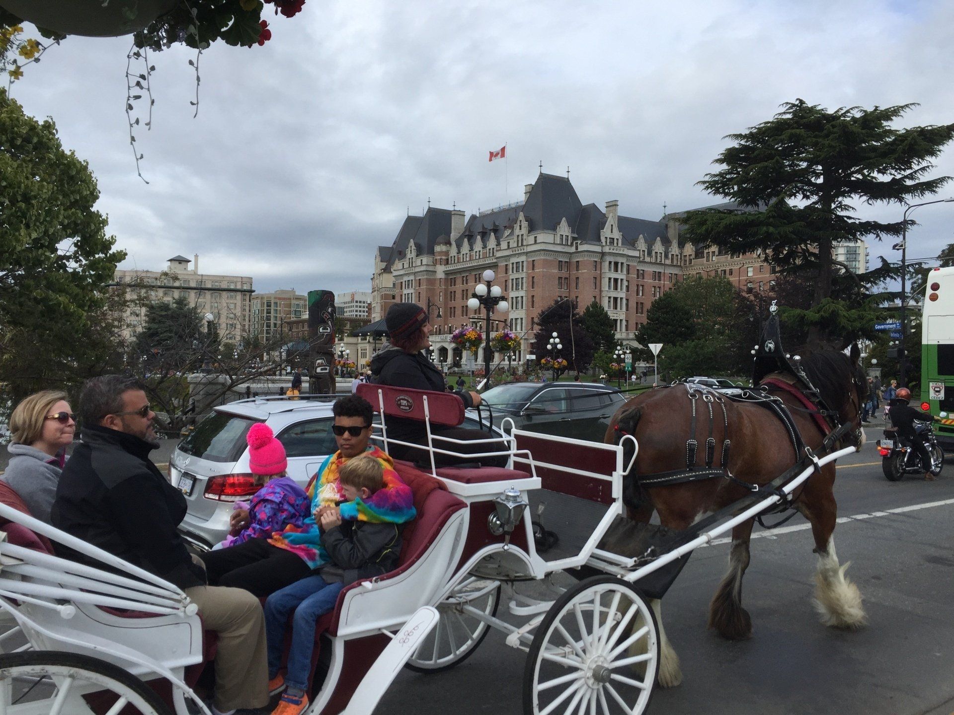 Horse-drawn carriage carrying passengers in front of a large brick building; cloudy sky.