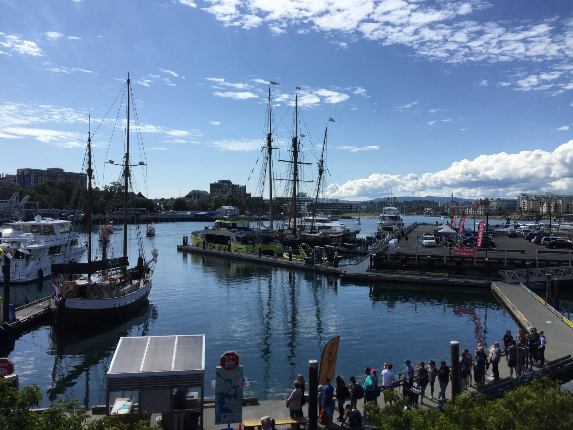 Harbor scene with sailboats, boats, docks, and a crowd of people under a bright, cloudy sky.